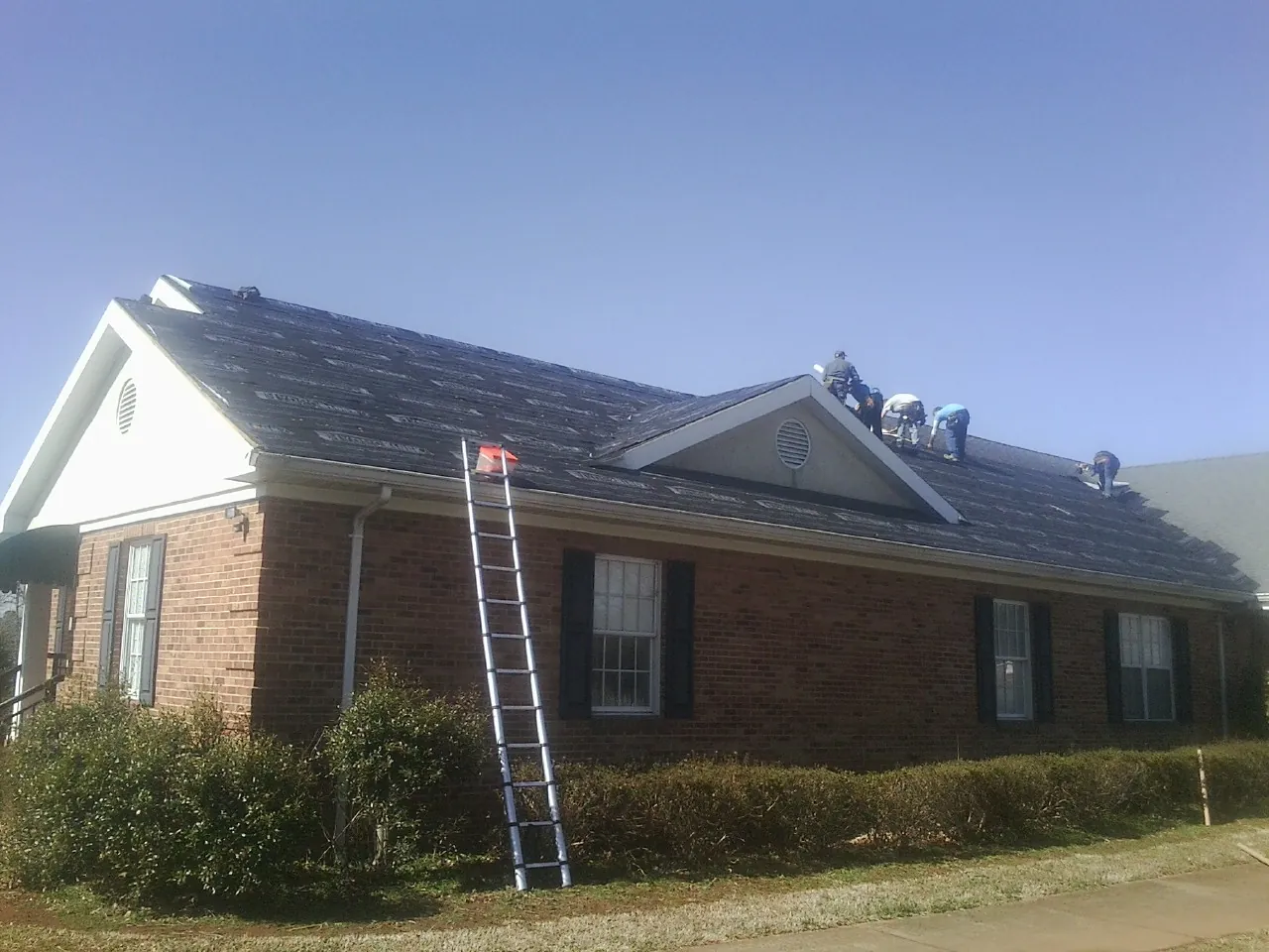 Crew tearing shingles off a brick ranch roof with a ladder set — Emergency Roof Repair work by our crew in Maxbass