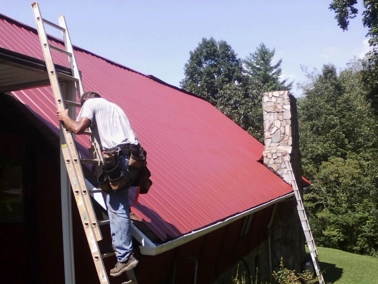 Roofer on a ladder beside a red metal roof and stone chimney — Soffit & Fascia Repair by Roofing Brothers in Maxbass